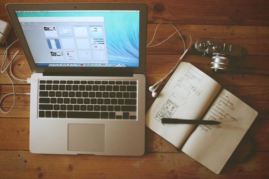 Ordinateur portable ouvert sur une table en bois, avec un carnet de croquis et un appareil photo